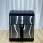 Stainless steel air fryer with black accents on a wooden surface and white curtain background
